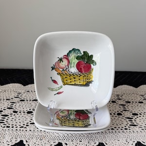 May include: Two white square ceramic plates with rounded corners. Each plate features a colorful illustration of a basket filled with vegetables, including tomatoes, carrots, and lettuce. The plates are displayed on a clear plastic stand.