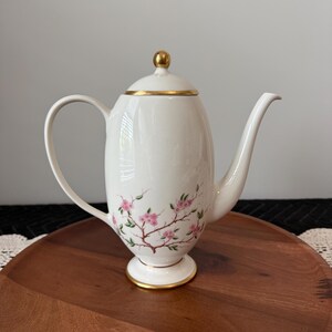 May include: A cream-colored porcelain coffee pot featuring a delicate floral pattern of pink blossoms on brown branches. The pot has a tall, slender body, a curved handle, a long spout, and a lid topped with a small, polished gold-tone knob.