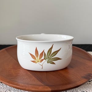 May include: White ceramic bowl with a handle, decorated with two autumn-colored maple leaves. The bowl is set on a round wooden tray. The leaves are in shades of brown, orange, and green.