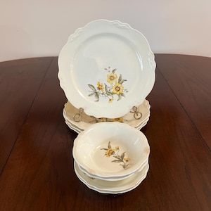 May include: Set of white ceramic dinnerware with a yellow floral pattern. The set includes a dinner plate, a salad plate, and a bowl.