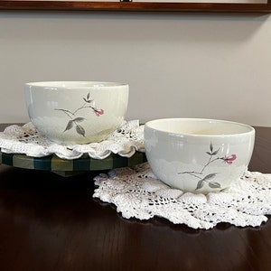 May include: Two white ceramic bowls with a floral design featuring pink flowers and gray leaves. The bowls are sitting on white crocheted doilies.