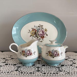 May include: A vintage porcelain tea set featuring a serving platter, creamer, and sugar bowl. The set has a light blue border and a floral design with pink, yellow, and purple roses. The pieces are arranged on a white lace tablecloth.