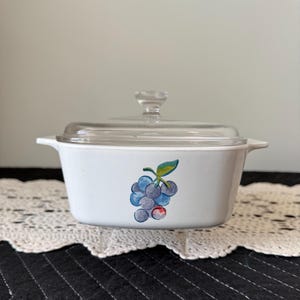 May include: White casserole dish with a clear glass lid. The dish features a painted design of blue grapes with green leaves. The dish has two handles and is sitting on a white lace doily.