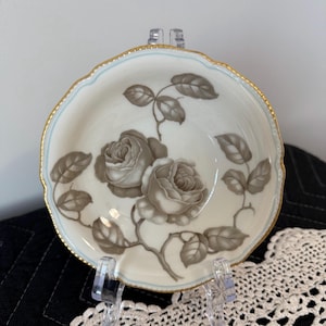 May include: A small, off-white dish with a floral rose design. The roses are a muted brown color, with brown leaves and stems. The rim has a gold trim with a light blue border. The dish is displayed on a clear stand.