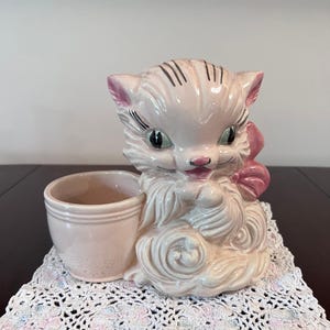 May include: A vintage ceramic planter in the shape of a pink cat with a bow. The cat has black stripes on its head and green eyes. The planter has a small, round opening for plants or other items. The planter is sitting on a white lace doily.