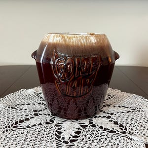 May include: A vintage brown ceramic cookie jar with a glossy finish. The jar has a rounded shape with a lighter brown top and the word "COOKIE" embossed on the front. Two small handles are on the sides.