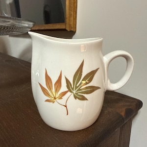 May include: White ceramic pitcher with a handle and spout. The pitcher features a design of two autumn-colored maple leaves. The leaves are in shades of brown, orange, and green. The pitcher is likely for serving beverages.