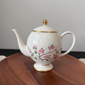 May include: A white ceramic teapot with a spherical body, a curved spout, and a handle. The teapot features a painted design of brown branches with pink blossoms and green leaves. It has a lid with a gold-colored knob and gold trim on the base.