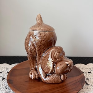 May include: A brown ceramic dog-shaped cookie jar with a lid. The jar is sitting on a wooden plate. The dog is depicted in a playful pose, with its rear end and tail forming the jar's body and its head and front paws at the front.