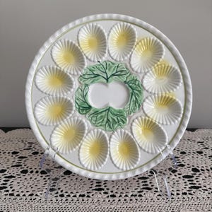 May include: White ceramic egg serving platter with 12 egg-shaped indentations, each with a yellow center. The platter has a green leaf design in the center and a rope-like border. The platter is displayed on a clear stand.