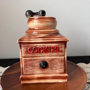 May include: A brown ceramic cookie jar with a lid and a black handle. The word "COOKIES" is written in red letters on the front. The jar has a drawer-like design and a black knob.