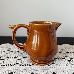 May include: A brown ceramic pitcher with a glossy finish. The pitcher has a curved handle and a spout for pouring. The base is round and the body has a bulbous shape. The pitcher is likely vintage and suitable for serving beverages.