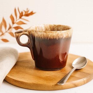 May include: A brown ceramic mug with a unique handle design, featuring a circular opening. The mug has a glossy finish and a lighter brown rim. A silver spoon rests on a wooden cutting board, next to a folded beige cloth.