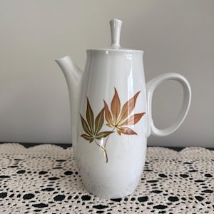 May include: White ceramic coffee pot with a lid and handle. The pot features a design of two maple leaves in shades of brown and green. The pot is sitting on a white lace tablecloth.
