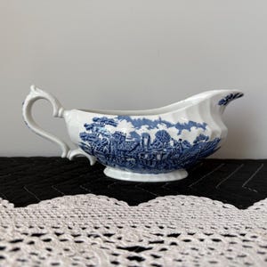 May include: White ceramic gravy boat with a blue and white landscape design. The gravy boat has a handle and a spout. The design features buildings, trees, and clouds. The gravy boat is on a black surface.
