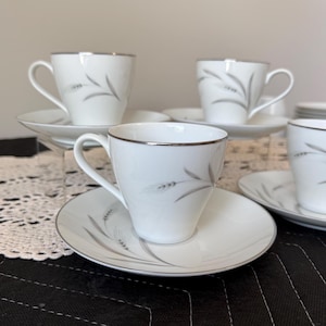 May include: A set of white porcelain teacups and saucers. Each cup and saucer features a silver rim and a gray wheat stalk design. The cups have a curved handle and the saucers are round.
