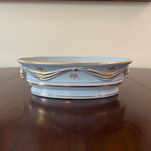 May include: A light blue porcelain serving dish with gold trim and floral accents. The oval-shaped dish features a decorative ribbon design with small pink and yellow flowers. The rim and base are also trimmed in gold.