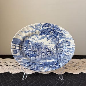 May include: A blue and white decorative plate with a scenic illustration. The plate features a detailed landscape with a horse-drawn carriage, buildings, and trees. The plate has a scalloped edge.