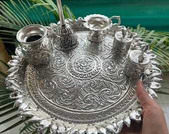 Puede incluir: Una bandeja de plata con intrincados diseños florales, que presenta una variedad de recipientes de plata y un diya de plata. La bandeja está decorada con un borde elevado de hojas estilizadas.