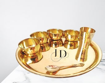 Puede incluir: Un juego de seis cuencos y una taza de latón dorado, dispuestos en un plato grande y redondo de latón dorado. El juego incluye una cuchara pequeña. El plato tiene el texto "JD Handicrafts" en negro.