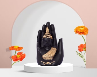 May include: Black Buddha statue with gold accents sitting in a black hand sculpture. The statue is on a white pedestal with orange flowers in the background.