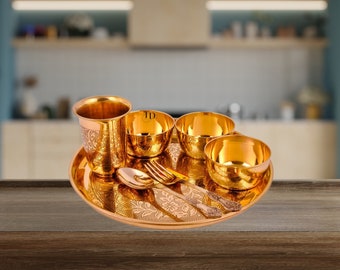 Puede incluir: Un juego de vajilla de metal dorado con un patrón floral decorativo. El juego incluye un plato, cuatro cuencos, un vaso, una cuchara y un tenedor.
