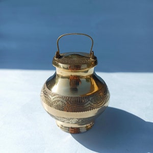 May include: A small, gold-coloured metal pot with a lid and a handle. The pot has an engraved floral pattern around the middle. The pot is sitting on a white surface with a blue background.