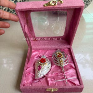 Puede incluir: Una caja de joyería de terciopelo rosa con tapa transparente, que contiene un colgante plateado ornamental en forma de lágrima con una rosa roja y una gema rosa, y una pluma plateada con detalles rojos y dorados. El texto "JAIDURGAHANDICRAFTS" es visible.
