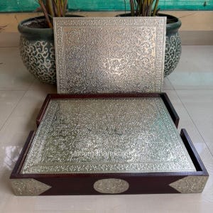 Puede incluir: Dos bandejas decorativas con superficies de metal plateado ornamentadas y marcos de madera marrón oscuro. Las bandejas presentan un detallado diseño floral. Una bandeja está dentro de un marco de madera, la otra descansa encima.