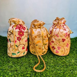 Puede incluir: Tres bolsas con cordón bordadas en beige con motivos florales en rojo, rosa y dorado. Cada bolsa tiene un cordón dorado y detalles de borlas. Las bolsas se exhiben sobre un lecho de hierba verde.