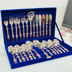 May include: A cutlery set presented in a blue velvet-lined case. The collection includes forks, spoons, and knives with polished silver-tone heads and textured, copper-coloured handles. Each piece is secured in individual slots within the box.