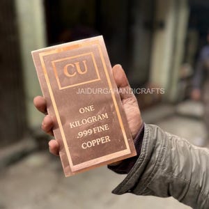 Może przedstawiać: Miedziany sztab z napisem "CU" w kwadracie oraz słowami "ONE KILOGRAM .999 FINE COPPER". Sztaba trzymana jest w dłoni osoby. Widoczny jest również tekst "JAIDURGAHANDICRAFTS".