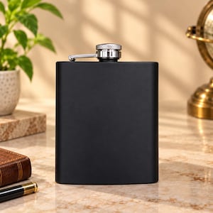 Puede incluir: Una petaca rectangular de color negro mate con cuerpo curvado y tapón de rosca de metal plateado. La petaca cuenta con un mecanismo de tapa con bisagra fijado en la parte superior, lo que proporciona un cierre seguro para el recipiente.