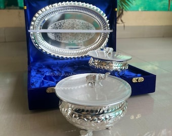May include: A silver-colored serving set with an oval tray and two lidded bowls, presented in a blue velvet box. The bowls feature elephant-shaped handles. The tray and bowls have ornate, engraved designs.