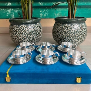 May include: A set of six silver teacups and saucers with ornate designs. The cups and saucers are arranged on a blue velvet box with gold accents.
