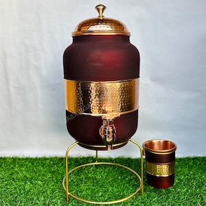 May include: A maroon water dispenser with a hammered gold band and lid, set on a gold stand. A matching cup sits beside it. The dispenser has a spigot for easy pouring. The overall height is approximately 20 inches.