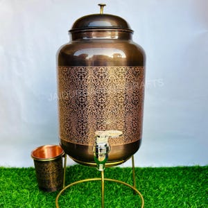 Puede incluir: Un dispensador de agua de color cobre con un patrón floral decorativo. El dispensador tiene un grifo y se apoya en un soporte dorado. Una taza de cobre a juego está al lado del dispensador. El dispensador tiene una tapa.