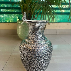 May include: A silver vase with intricate floral and leaf designs. The vase has a wide mouth and a narrow neck. A small silver bird figurine sits on the rim of the vase.