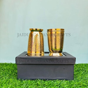 May include: Two gold-coloured metal drinking glasses with ornate floral patterns. The glasses are displayed on a black box, set against a backdrop of green grass and a light blue wall. The text "JAIDURGAHANDICRAFTS" is visible.