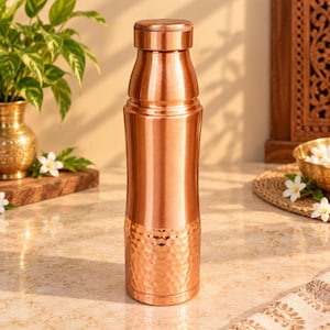 Puede incluir: Una botella de agua cilíndrica de cobre con una sección superior lisa y un acabado martillado texturizado en la mitad inferior. La botella cuenta con una tapa de rosca a juego con una parte superior plana y un cuello ligeramente cónico.