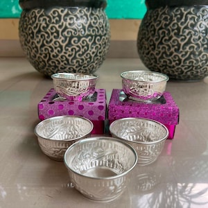 May include: Four silver bowls with intricate designs are displayed on pink boxes with polka dots and checkered patterns. The bowls are arranged in a staggered pattern, with two bowls on each box.