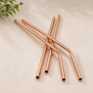 Puede incluir: Un juego de cuatro pajitas de metal en color oro rosa, que incluye dos pajitas rectas y dos pajitas dobladas. Las pajitas tienen un acabado liso y reflectante con forma cilíndrica. El texto jaidurga crafts es visible en el centro de la imagen.