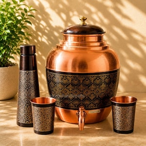 Puede incluir: Un dispensador de agua de cobre con tapa marrón oscura y una banda negra con patrón floral dorado, acompañado de una botella alta a juego y dos vasos. El dispensador tiene un grifo de metal y una pequeña etiqueta que dice Clazinox.