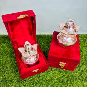 May include: Two silver decorative containers with lids and ornate patterns are shown. One is inside an open red velvet box, the other on a closed red velvet box. The boxes have gold-coloured clasps. The containers are likely for decorative purposes.
