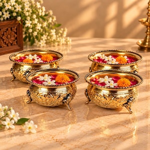Könnte beinhalten: Vier runde Messingschalen mit detaillierten Blumenverzierungen und drei kleinen Füßen. Jede Schale ist mit Wasser, leuchtend rosa Rosenblättern, weißen Jasminblüten und leuchtend orangefarbenen Ringelblumen gefüllt, was ein dekoratives Arrangement ergibt.