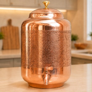Könnte beinhalten: Ein zylindrischer Wasserspender aus Kupfer mit polierter Oberfläche, einem Mittelband mit eingeprägtem Blumenmuster, einem passenden Deckel mit kleinem goldfarbenen Knauf und einem funktionalen Kupferhahn an der Vorderseite.