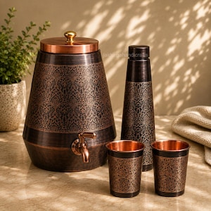 Könnte beinhalten: Ein Wasserspender aus Kupfer mit Deckel und Zapfhahn, eine hohe Wasserflasche und zwei passende Becher. Jeder Gegenstand hat ein dunkles, gebürstetes Finish mit einem komplizierten, sich wiederholenden Blumenmuster in hellerem Kupferton.