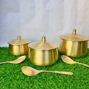 Peut inclure: Un ensemble de trois récipients en laiton avec couvercles et deux cuillères. Les récipients sont de tailles différentes, de forme arrondie et avec un petit bouton sur chaque couvercle. Les cuillères ont un design simple. Sur un fond vert.