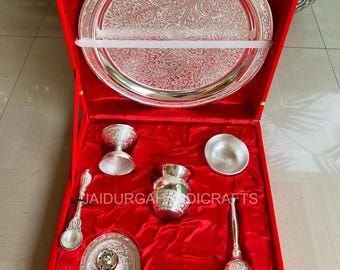 May include: A silver-colored puja set displayed in a red velvet-lined box. The set includes a large, intricately patterned tray, a small cup, a spoon, a small pot, a plate, a bell, and a small bowl. The text "JAIDURGA DICRAFTS" is visible.