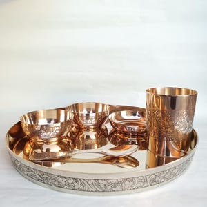 May include: A set of copper-toned bowls and a cup with intricate floral designs. The bowls and cup are arranged on a large, round copper-toned plate with a silver-toned border. The plate has a decorative floral design around the edge.